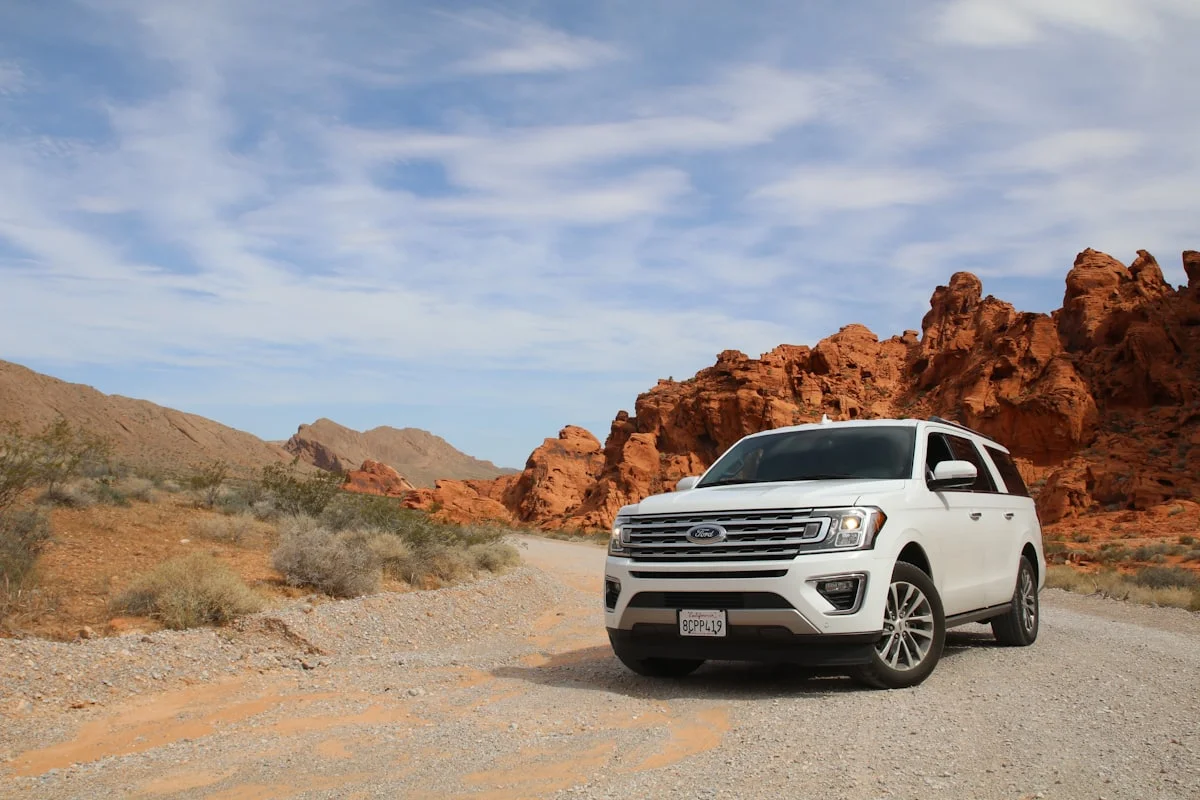 White Ford Expedition seven-seater parked on a gravel desert track beside red sandstone outcrops.